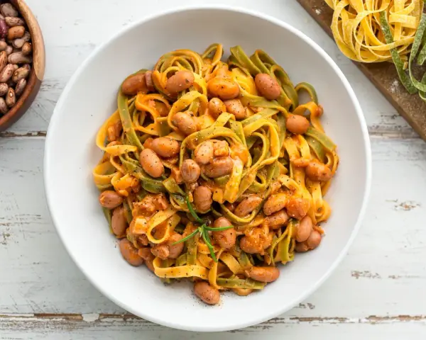 Pasta all'uovo con fagioli borlotti in una ciotola bianca, con fagioli secchi e pasta fresca colorata sullo sfondo.