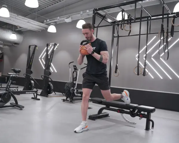 Man utför rumplyft på gym med kettlebell. Träningsredskap som roddmaskiner och ringar syns i bakgrunden.