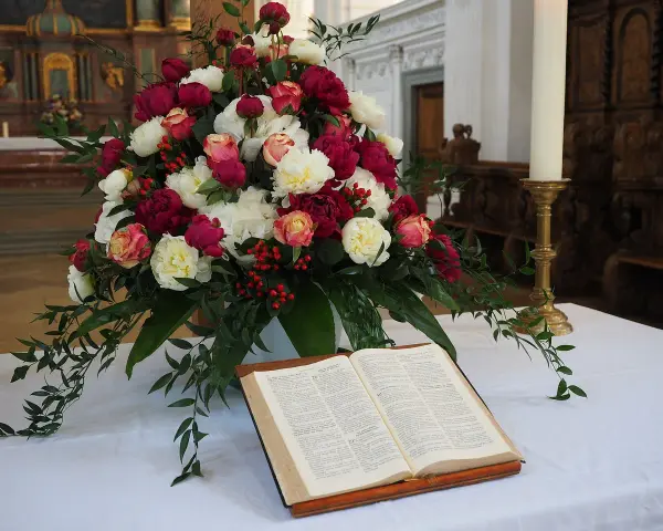 Piękne bukiety do kościoła, florystyka przed ołtarzem. Kompozycja z róż i piwonii, obok otwarta księga i świeca.