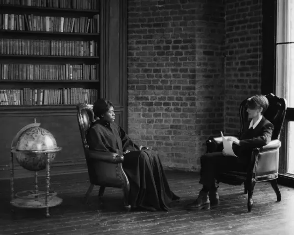Deux personnes assises dans des fauteuils en cuir, une femme en robe longue et un homme en costume, discutent dans une bibliothèque. Scène évoquant des témoignages procédure en départage prud'hommes.