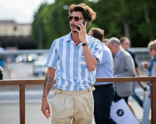Hombre con camisa a rayas azules y blancas y tiro pantalón alto color beige, hablando por teléfono.
