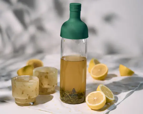 Orzeźwiający cold brew tea w butelce z zieloną zakrętką, obok szklanki z lodem i cytryny. Idealny na upalne dni.