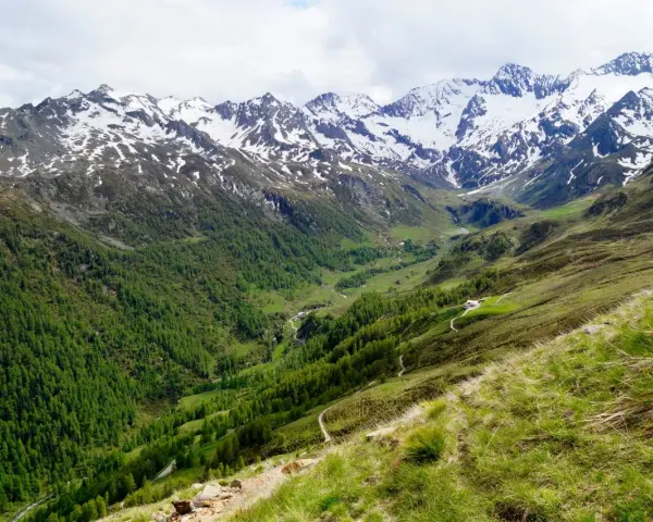 Un sentiero serpeggia su un pendio erboso verso un passo rombo in moto, con montagne innevate sullo sfondo.