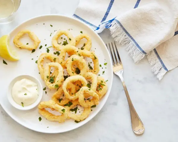 Crujientes calamares a la romana, servidos con limón y salsa tártara en un plato blanco.