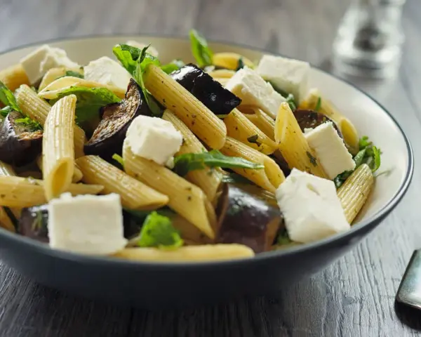 Piatto di pasta con melanzane in bianco, cubetti di formaggio fresco e foglie verdi.