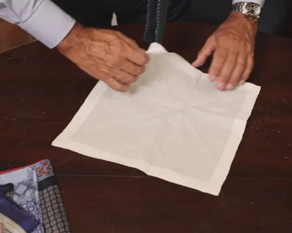 Mani esperte mostrano come piegare fazzoletto da taschino su un tavolo di legno. Altri fazzoletti colorati sono visibili a sinistra.