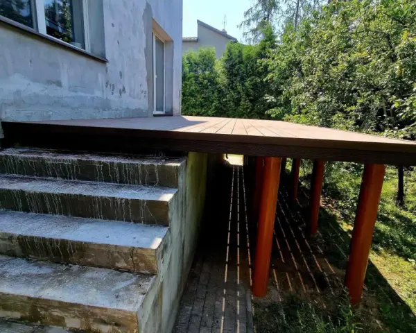 Nowy, wysoki taras na słupach, z drewnianym blatem, sąsiaduje z betonowymi schodami i zielonym ogrodem.