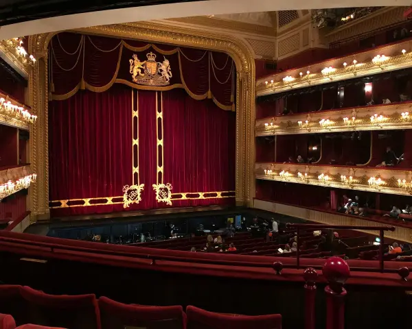 Ein prunkvolles Opernhaus mit roten Samtvorhängen und goldenen Verzierungen. Die Logen sind mit Lichtern geschmückt, und im Parkett sitzen Zuschauer.