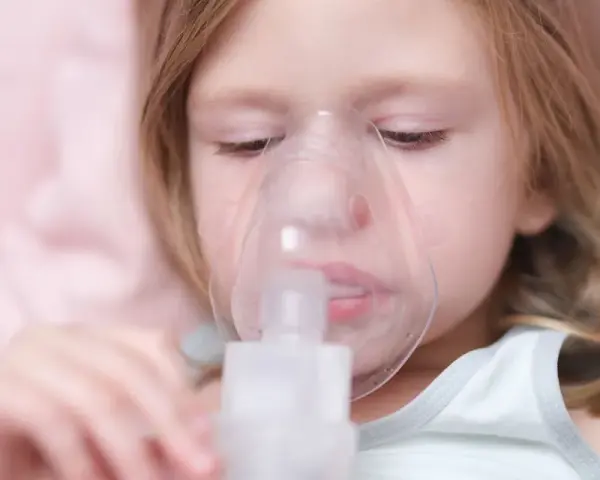 Mała dziewczynka z maską inhalacyjną na twarzy. Dziecko przechodzi inhalację na kaszel, by poczuć ulgę.