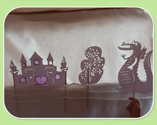 Théâtre d'ombres : château, arbre et dragon en papier, manipulés par une main.