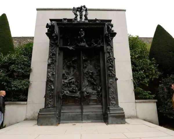 La Porte de l'Enfer de Rodin, une œuvre monumentale en bronze, se dresse au milieu d'un jardin. Deux visiteurs contemplent cette sculpture complexe.
