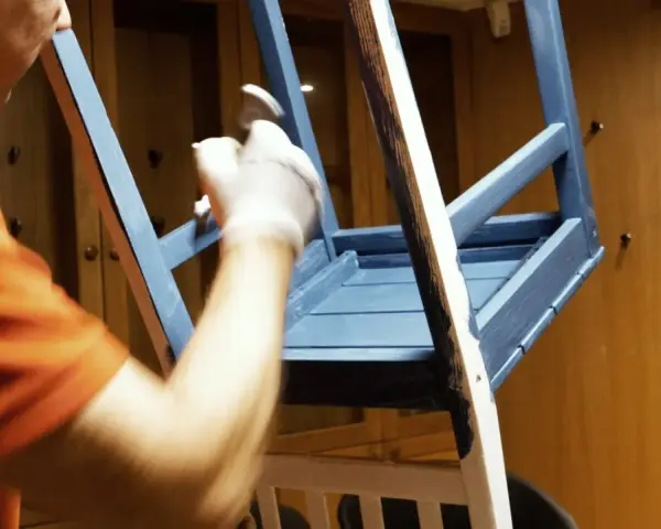 Person i orange tröja målar en blå stol med pensel.