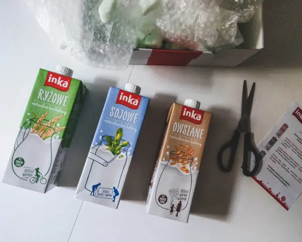 Trzy kartony napojów roślinnych Inka: ryżowy, sojowy i owsiany. Obok nożyczki i ulotka. Tak powstaje mleko owsiane.