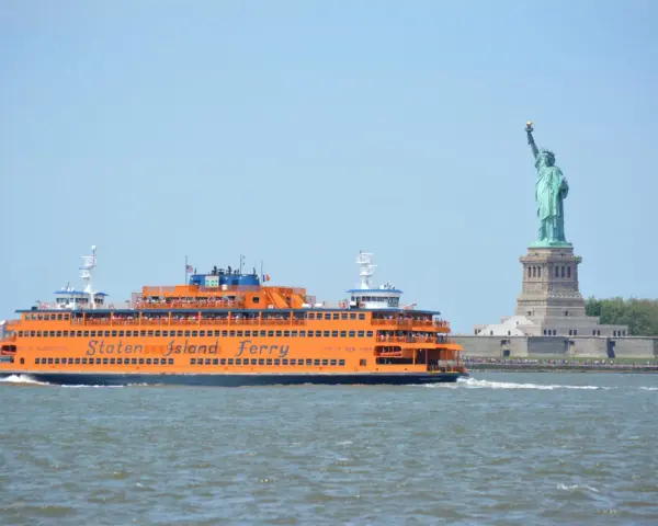 Pomarańczowy prom Staten Island Ferry płynie obok Statuy Wolności.