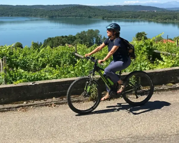 Donna in bici elettrica lungo il cammino di Oropa, tra vigneti e un lago sullo sfondo.