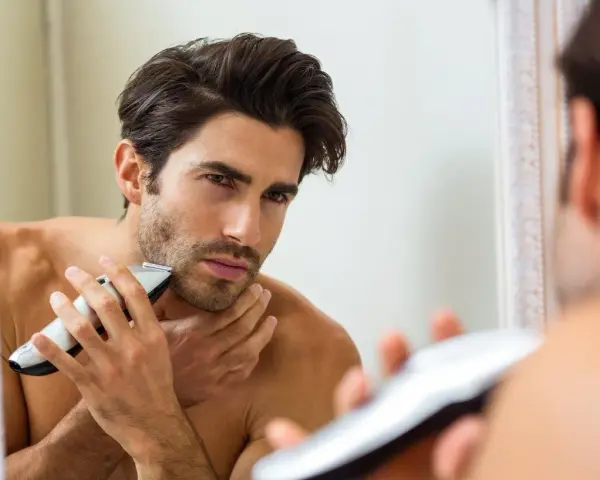 Hombre se afeita a contrapelo con una recortadora, mirándose en el espejo.