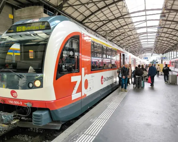 A train labeled "PE Interlaken, Ost" waits at the station, ready for its journey on the Lucerne to Interlaken Golden Pass line. Passengers gather on the platform.
