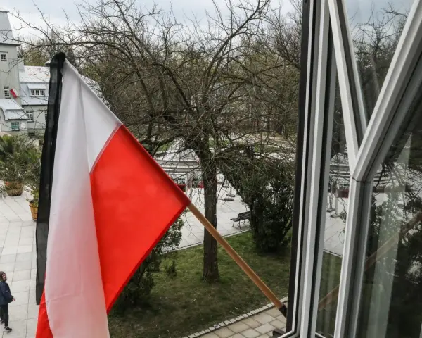 Flaga państwowa w biało-czerwonych barwach powiewa na wietrze. W tle widać drzewa i budynki.