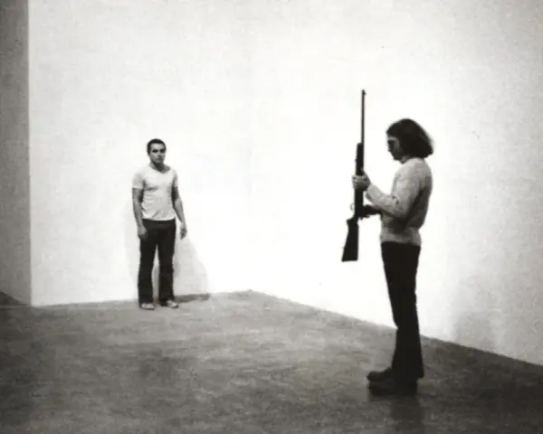 Chris Burden, dans une performance tendue, tient un fusil. Un homme se tient à distance, le regard fixé.