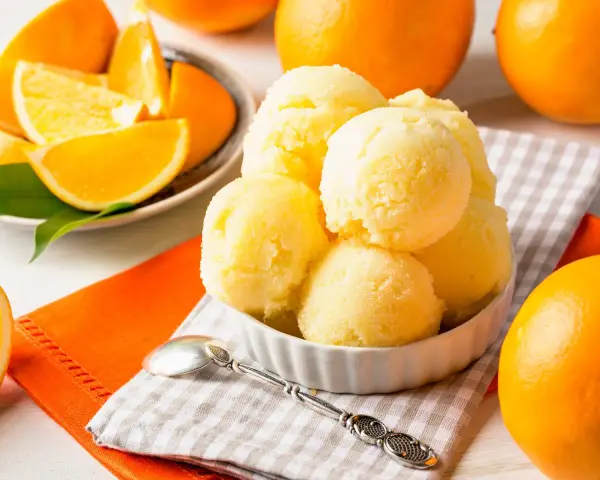Erfrischendes orangen sorbet in einer Schale, umgeben von frischen Orangen und saftigen Orangenstücken. Ein Löffel liegt bereit.