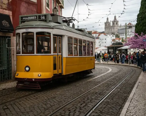 Żółty tramwaj 28 na brukowanej ulicy w Lizbonie, z ludźmi czekającymi na przystanku i kościołem w tle.