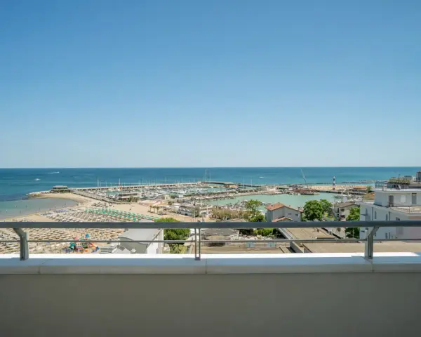 Vista dal balcone dell'hotel Acropolis Cattolica: spiaggia, porto turistico e mare azzurro sotto un cielo limpido.
