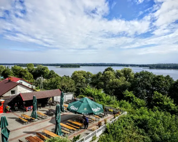 Malowniczy widok na park krajobrazowy pojezierza iławskiego. Taras z zielonymi parasolami i ławkami, jezioro z żaglówką w tle.