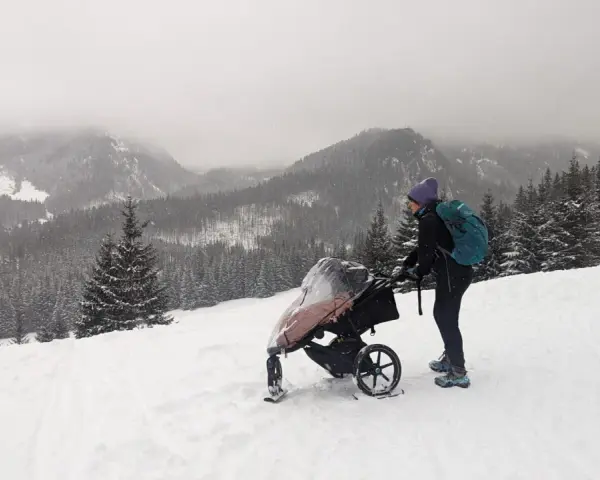 Zimowe Tatry z wózkiem. Kobieta w czapce i z plecakiem pcha wózek po ośnieżonym stoku, podziwiając górski krajobraz.