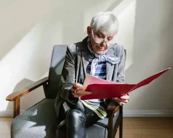 Une femme âgée, en pleine retraite, examine des documents artistiques dans un dossier rouge.