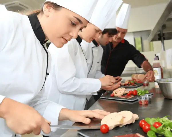 Des chefs apprennent la réglementation atelier cuisine en préparant des mets. Ils coupent du foie gras, des tomates cerises et de la salade.