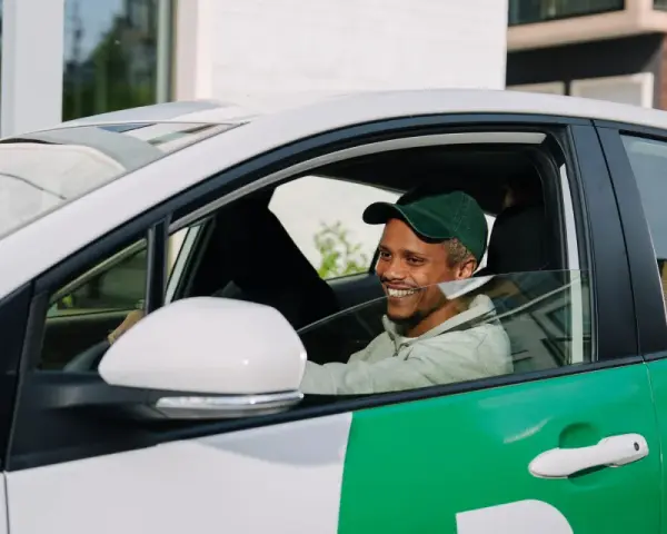 Lächelnder Mann mit grüner Kappe am Steuer eines weißen Autos mit grünen Bolt-Aufklebern. Er wird bald ein Bolt-Fahrer sein.