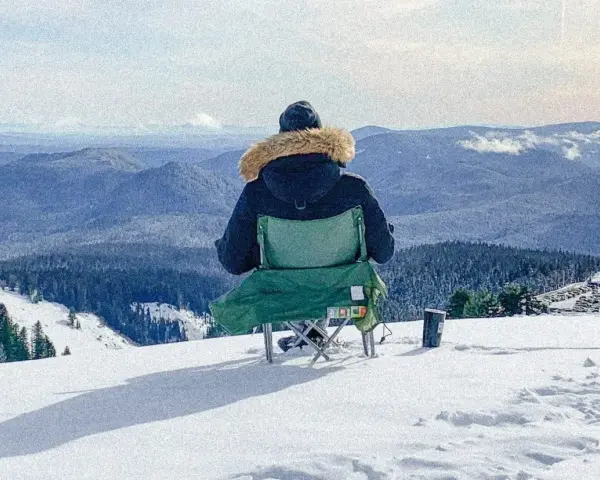 Persona sentada en silla plegable en la nieve, mirando un paisaje montañoso cubierto de árboles.