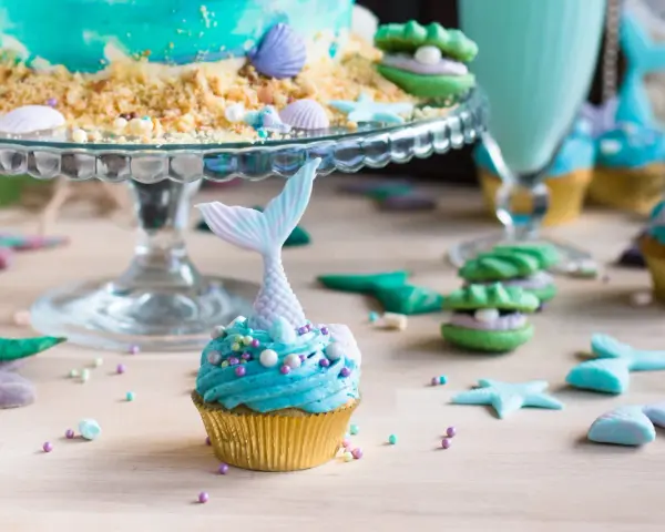 Meerjungfrau Muffins mit blauer Glasur und Schwanz-Dekorationen, umgeben von Muscheln und Seesternen.