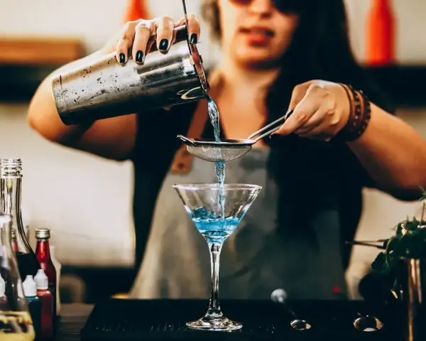 Barman przygotowuje drinka, przelewając niebieski płyn przez sitko do kieliszka. W tle widać butelki z różnymi nazwami drinków alkoholowych.