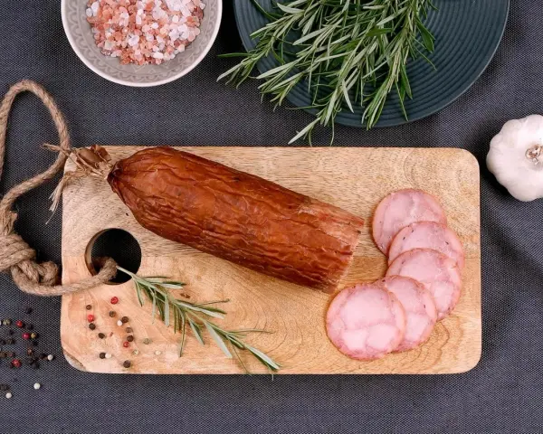 Kiełbasa żywiecka na desce, obok gałązka rozmarynu, czosnek i kolorowy pieprz. Kilka plastrów już pokrojonych.