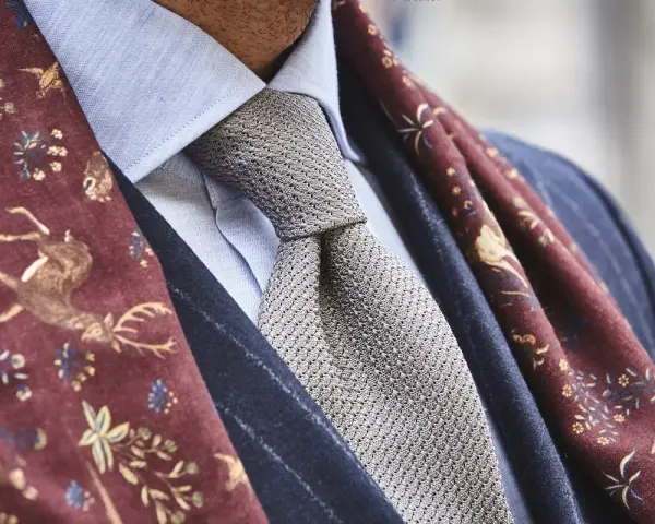 Elegancki mężczyzna w błękitnej koszuli, szarym krawacie i bordowym szalu z motywem jeleni. Całość emanuje nonszalancką elegancją, czyli sprezzaturą.