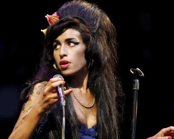 Amy Winehouse z charakterystyczną fryzurą i makijażem, śpiewająca do mikrofonu. Jej styl przypomina legendy z klubu 27, a jej muzyka wciąż żyje w sercach fanów w Polsce.