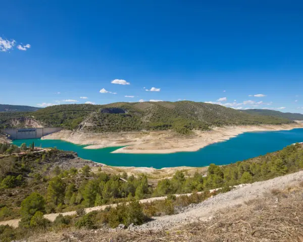 El pantano de Sacedón, con su agua turquesa, se extiende entre colinas verdes bajo un cielo azul.