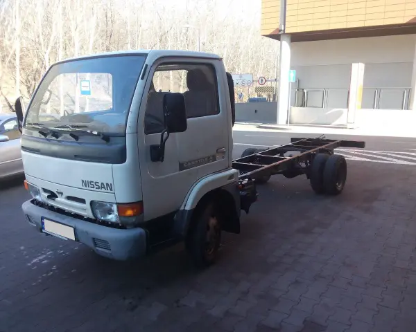 Biały Nissan Cabstar bez zabudowy, z widocznym gołym podwoziem i podwójnymi tylnymi kołami, stoi na kostce brukowej.