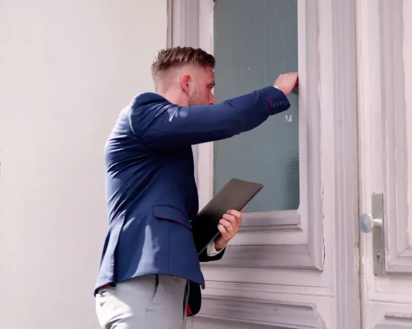 Mężczyzna w granatowej marynarce puka do drzwi, trzymając teczkę. Wygląda jakby prowadził skip tracing smartney.