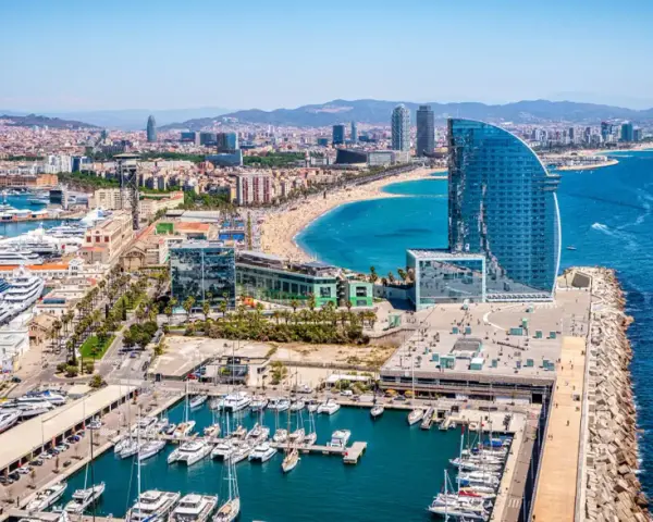 Barcelona morze: luksusowe jachty w marinie, plaża pełna ludzi i nowoczesny hotel w kształcie żagla.