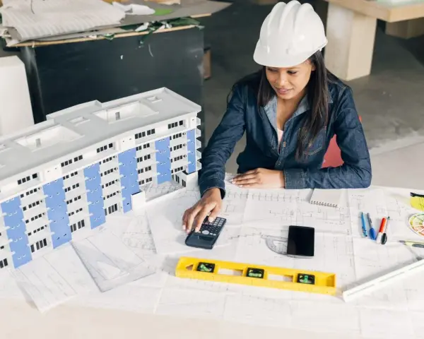 Architektka w kasku analizuje projekt budynku, używając kalkulatora. To może być związane z bms co to jest za system.