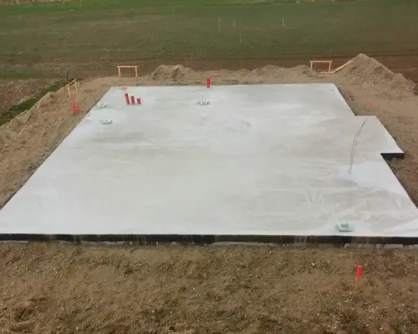 Płyta fundamentowa koszt budowy domu. Betonowa płyta fundamentowa z wystającymi rurami i elementami instalacji, otoczona ziemią i żwirem.
