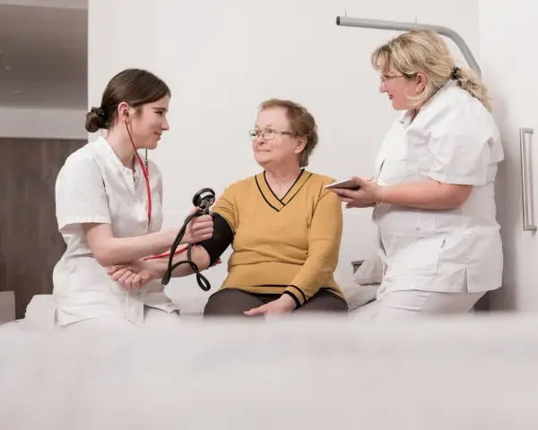 Zwei Pflegekräfte messen einer älteren Frau den Blutdruck. Ein Einblick in die Praxis des Rehabilitationspsychologie Studiums.