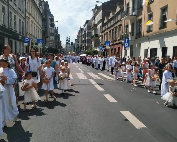 Procesja dzieci w białych strojach, niosących koszyki, idzie ulicą. W tle widać budynki i ludzi, co może symbolizować sekularyzację społeczeństwa.