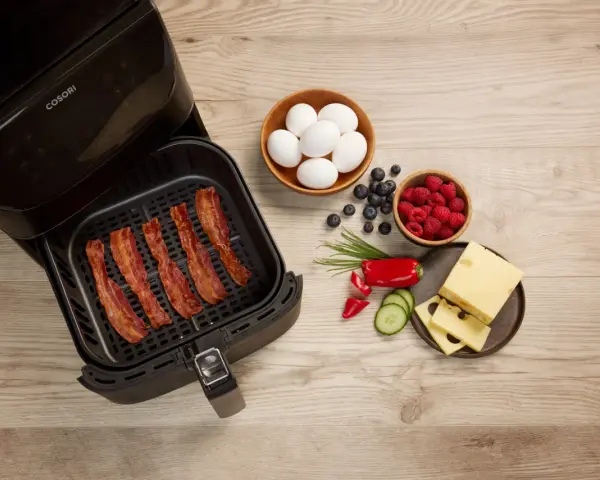 Knuspriger Bacon im Airfryer, daneben Eier, Beeren, Käse und Gemüse für ein perfektes Frühstück.