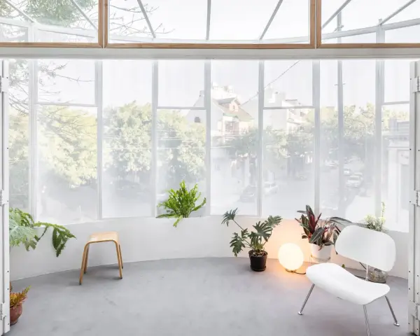 Interno luminoso con piante, sedia bianca e sgabello in legno, affacciato su una strada alberata da una veranda balcone.