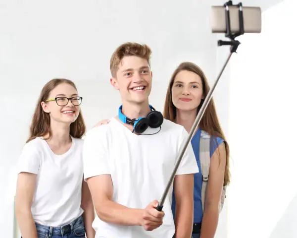 Trzech przyjaciół robi selfie na kijku. Chłopak z przodu uśmiecha się szeroko, dziewczyna w okularach po lewej stronie też, a ta z tyłu patrzy w obiektyw. To świetny sposób, jak robic dobre zdjecia!