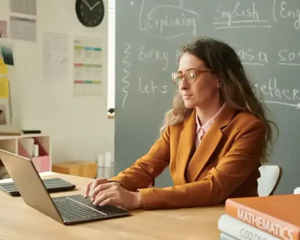 Nauczyciel początkujący w okularach pracuje przy laptopie w klasie. Na tablicy napisy po angielsku.