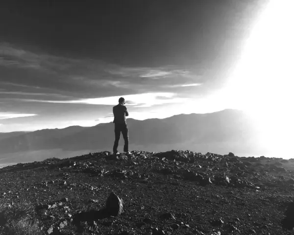 Ein Mann steht auf einem felsigen Gipfel und blickt in die Ferne. Die Sonne bricht durch die Wolken und beleuchtet die Berge. Vielleicht fragt er sich, was ist Weisheit.